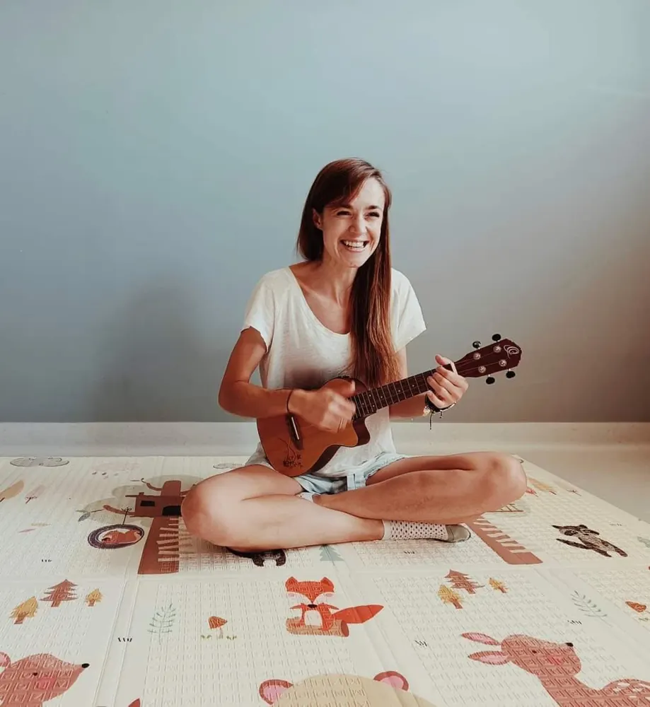 Prowadząca zajęcia umuzykalniające gra na ukulele, siedząc na kolorowej macie